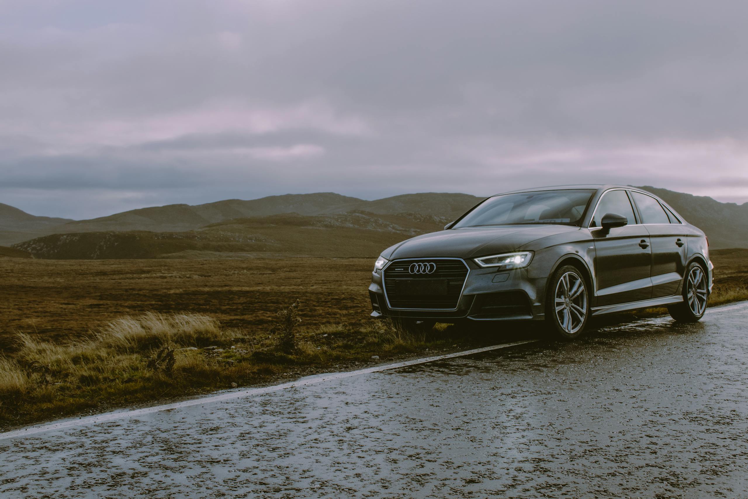 A luxury Audi sedan parked on a wet mountain road with moody skies and a scenic backdrop.