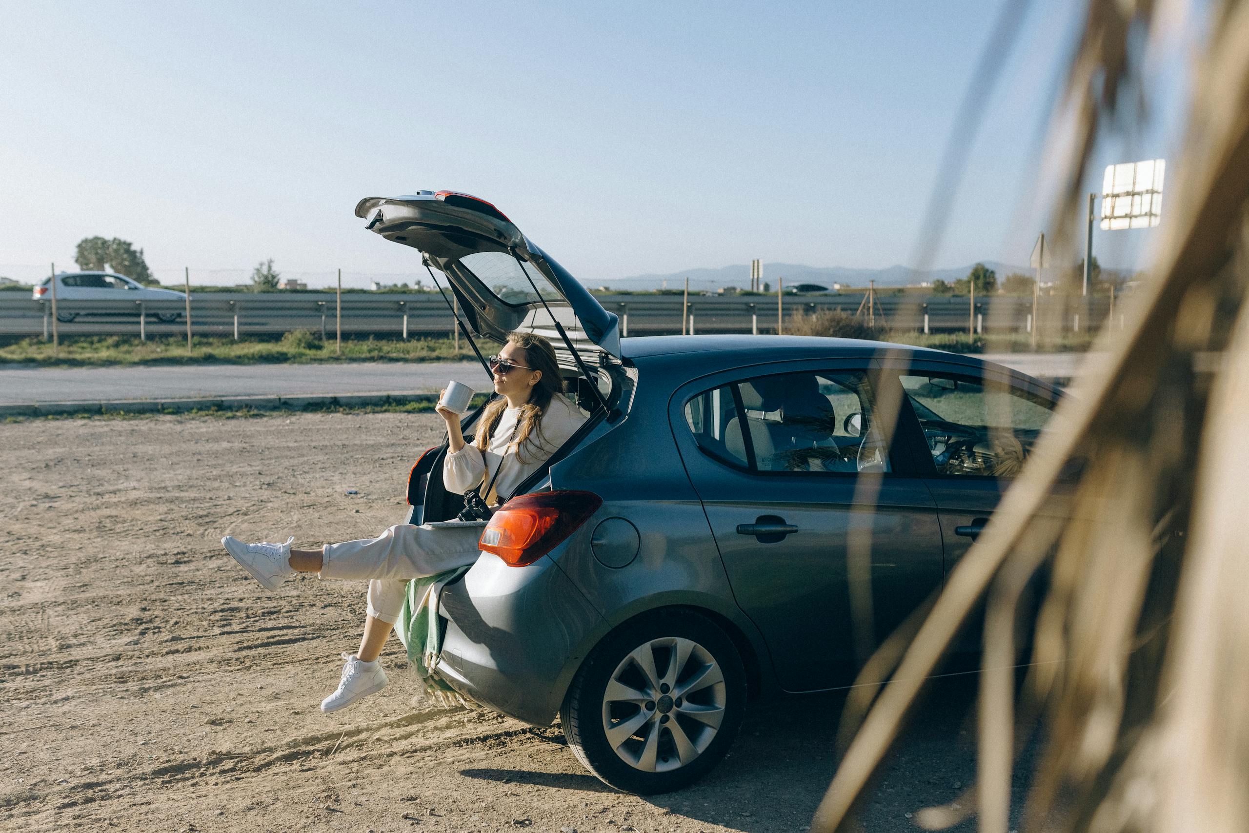 A woman sitting in the trunk of a parked car on a sunny day, enjoying a break during a road trip.