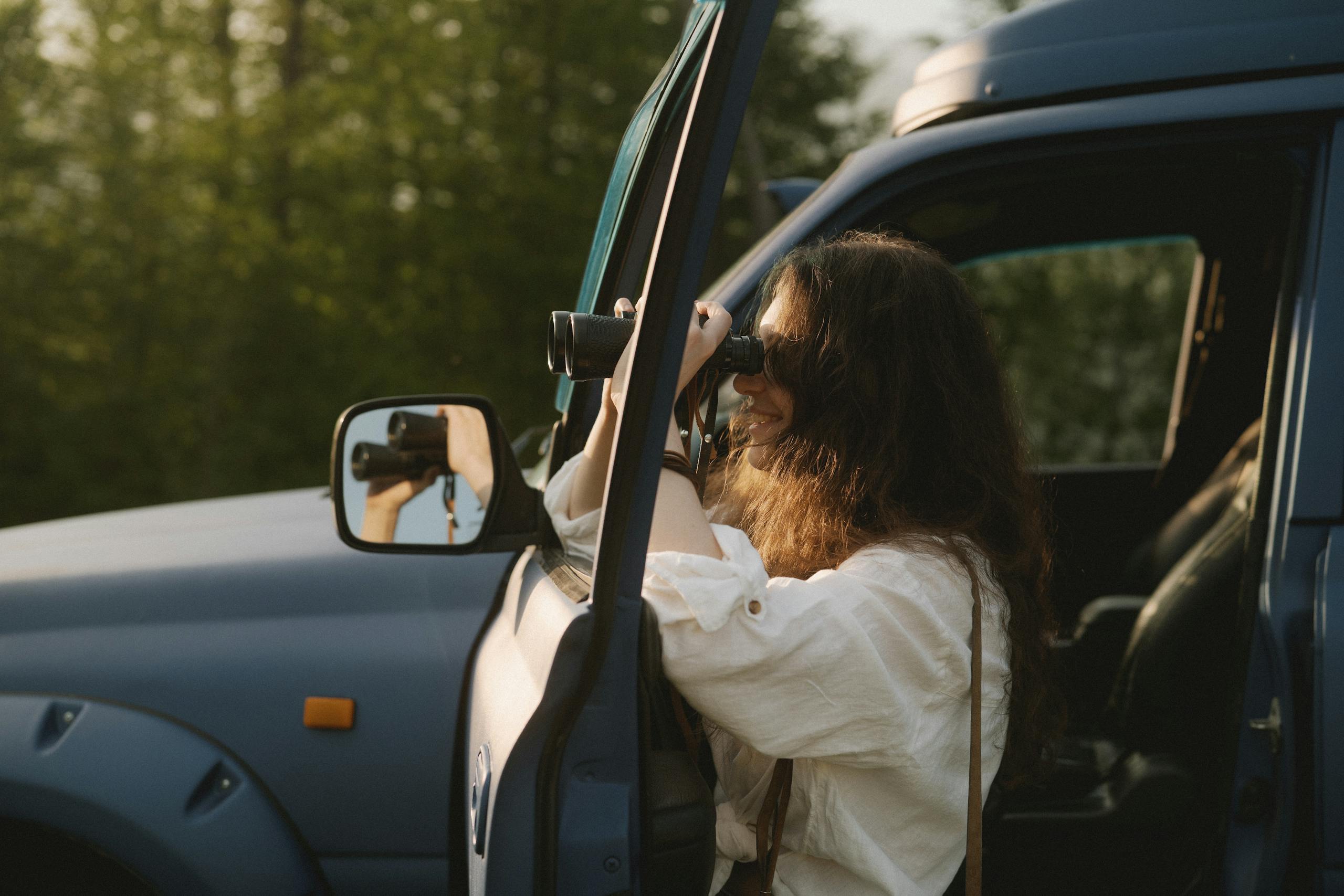 A woman using binoculars to observe nature from her car during a sunny day.